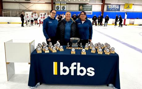 BFBS personnel Gini, Melissa stood in front of a table with trophies on. Ice hockey players in background.